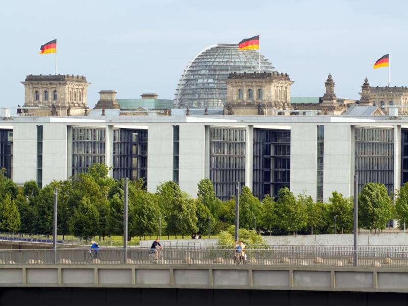 Kanzleramt und Bundestag, davor Bäume und Spreebrücke mit Radfahrenden